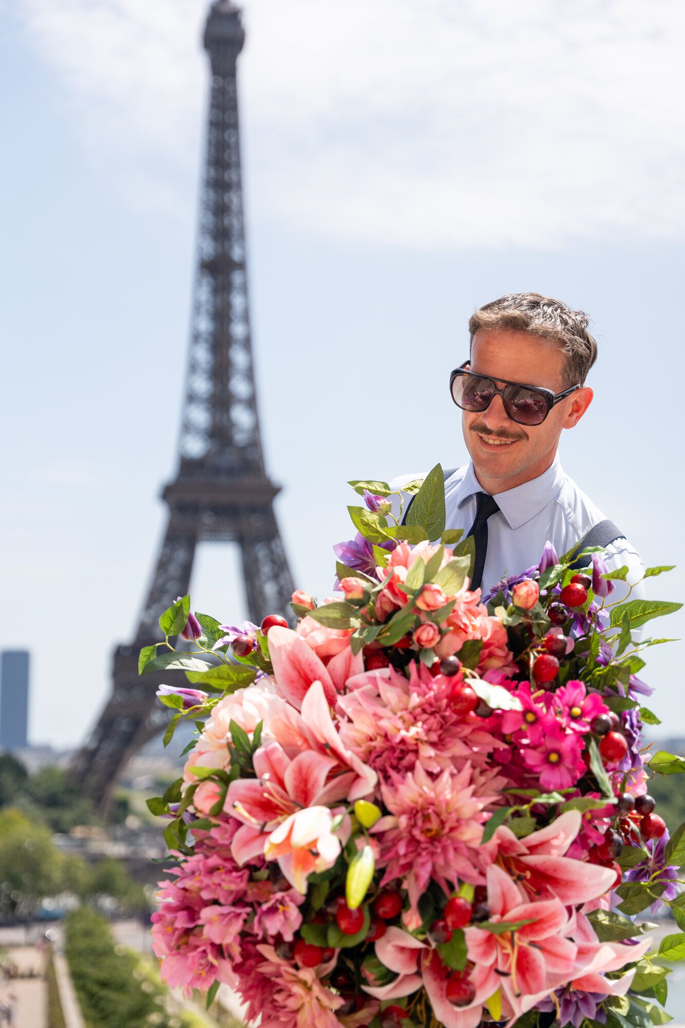 Sébastien Chol tenant une brassée de lys et de fleurs roses devant la tour Eiffel